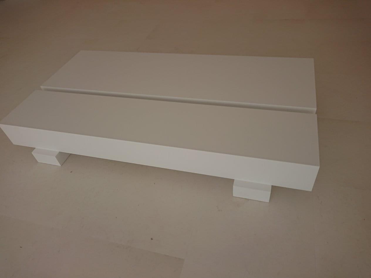 Dwa białe, lakierowane blaty drewniane z prostokątnymi wspornikami, leżące na płaskiej powierzchni, widoczne z boku.