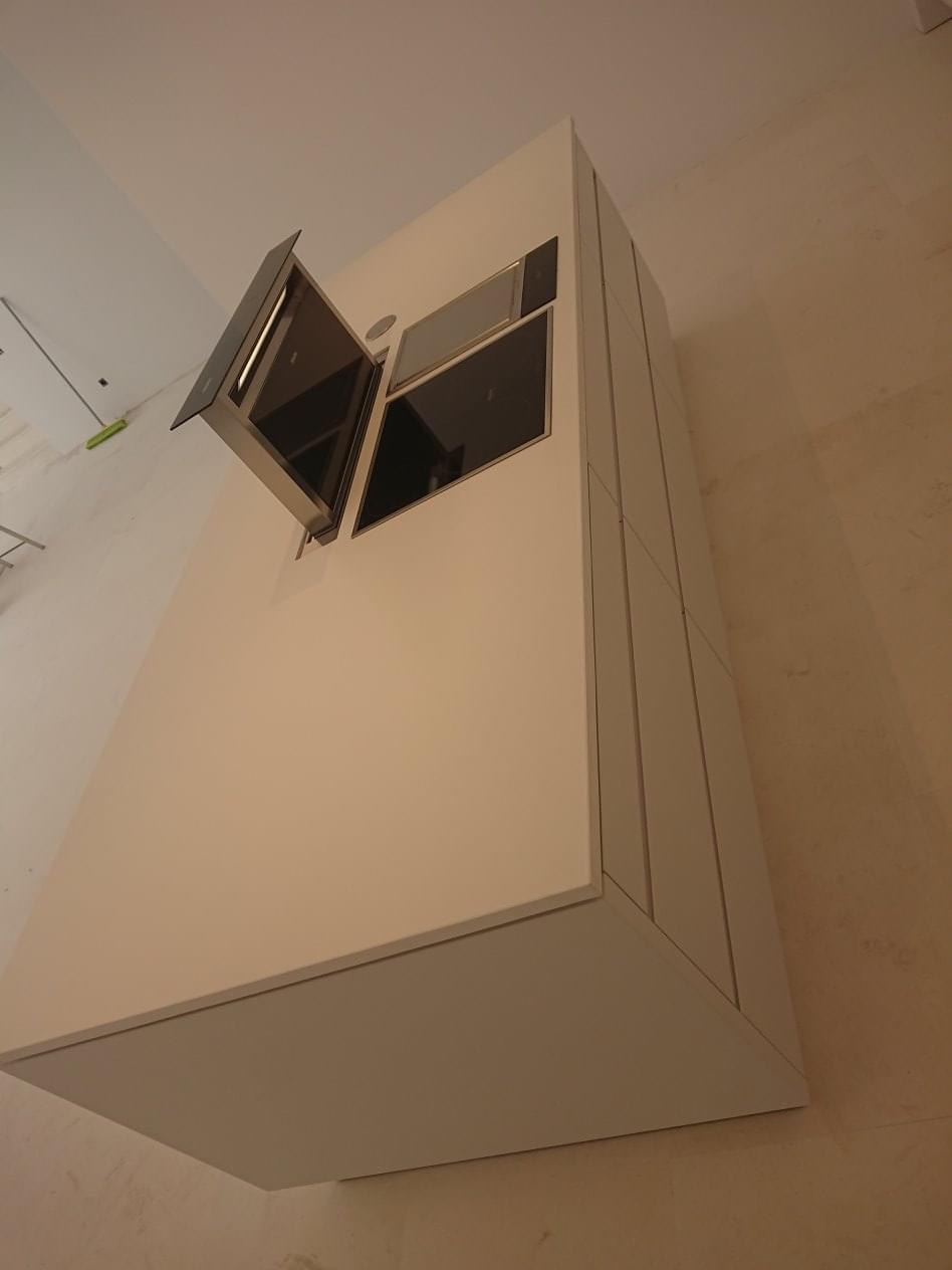 Nowoczesna, biała zabudowa kuchenna z wbudowanymi piekarnikami i otwartym okapem, widok z dołu na tle jasnej ściany.