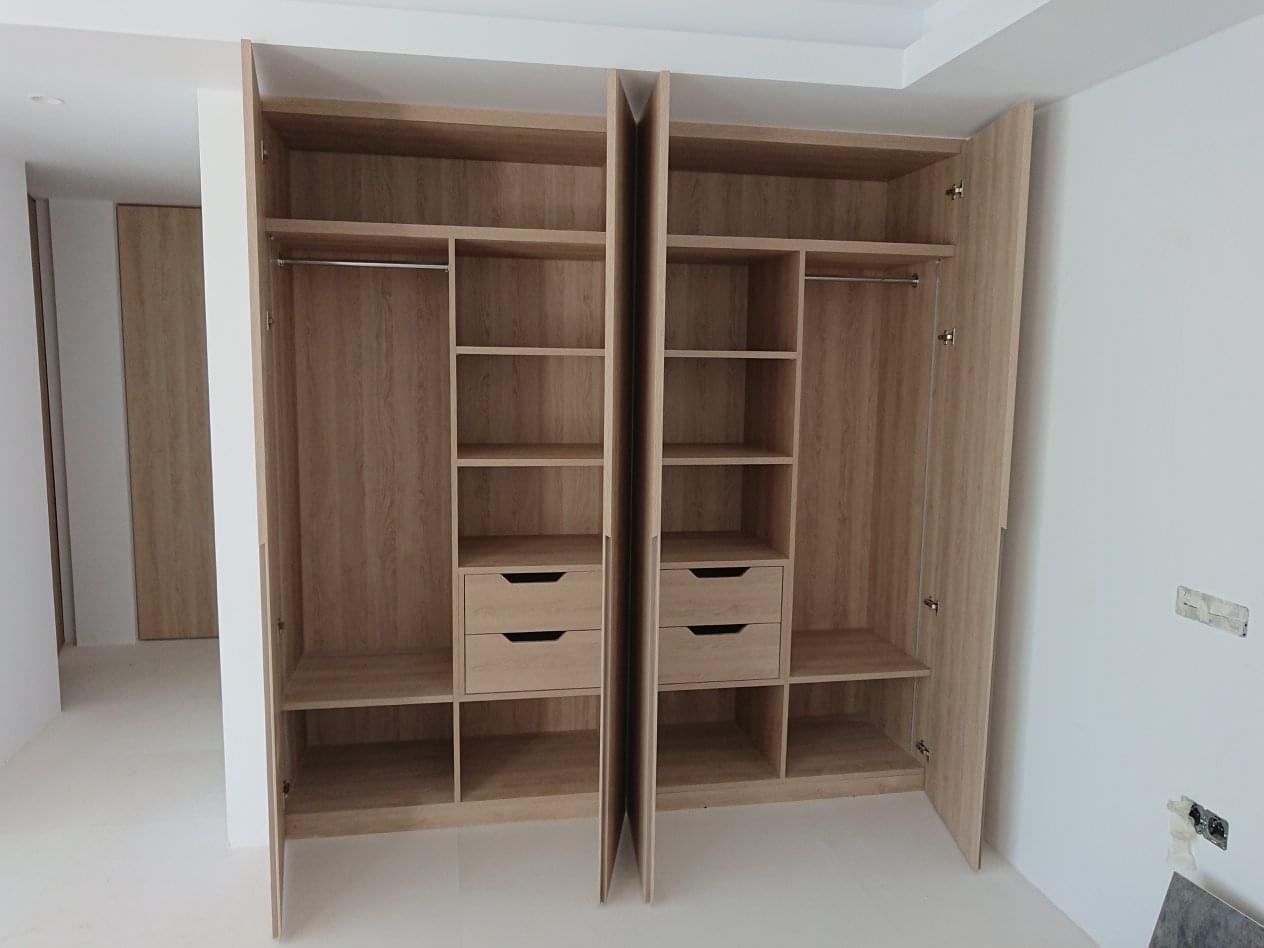 Dwie otwarte szafy z jasnego drewna z półkami, drążkiem i szufladami, widoczne w surowym wnętrzu, z niedokończonymi ścianami i podłogą.