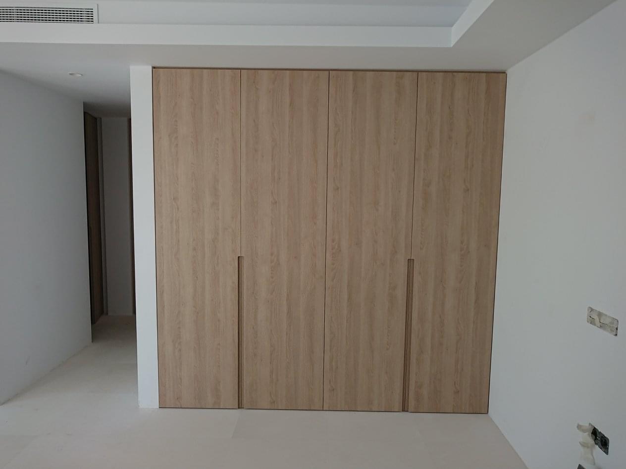 Czterodrzwiowa szafa wnękowa z jasnego drewna o widocznej strukturze usłojenia, z minimalistycznymi, pionowymi uchwytami, w jasnym pomieszczeniu z widoczną wentylacją na suficie.