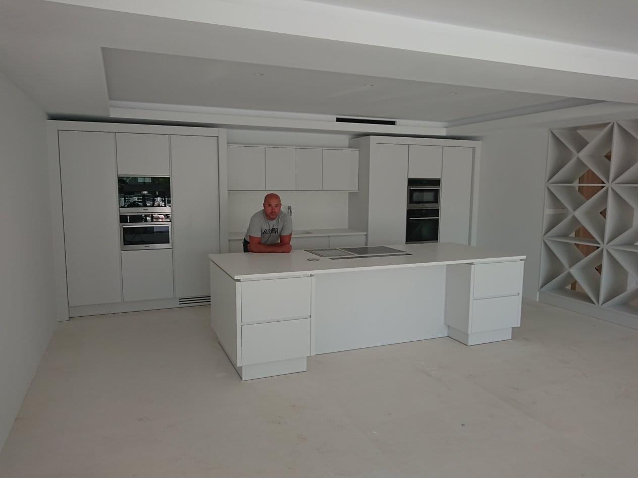 Jasna, minimalistyczna kuchnia z wyspą i zabudową pod sufit. Mężczyzna opiera się o wyspę. Widoczny fragment ażurowej ściany.