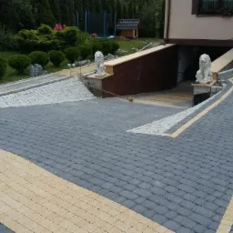 Ułożona z kostki brukowej dwukolorowa podjazd do garażu z dekoracyjnymi figurami lwów, mężczyzna w tle zamiata powierzchnię.