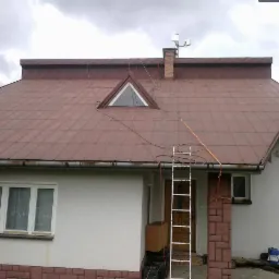 Dom jednorodzinny z brązowym dachem blaszanym, drabiny opierają się o rynnę i dach, widoczny komin z antenami oraz trójkątne okno dachowe.