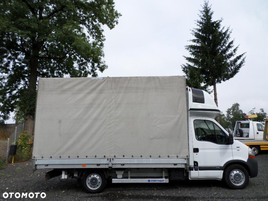 Biały samochód dostawczy z plandeką na tle drzew i nieba. Widoczny z boku, z logo 'Partner' na progu. W tle inny pojazd. Pochmurny dzień.