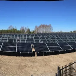 Rozległa farma paneli słonecznych w trakcie montażu, widoczne metalowe konstrukcje nośne, teren piaszczysty, w tle las i błękitne niebo.