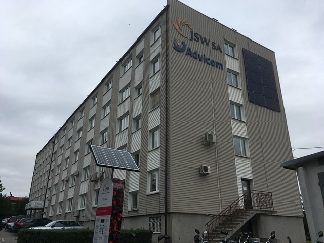 Budynek biurowy z zamontowanymi panelami słonecznymi na elewacji i wolnostojącym panelem z ładowarką, logo JSW SA i Advicom na ścianie.