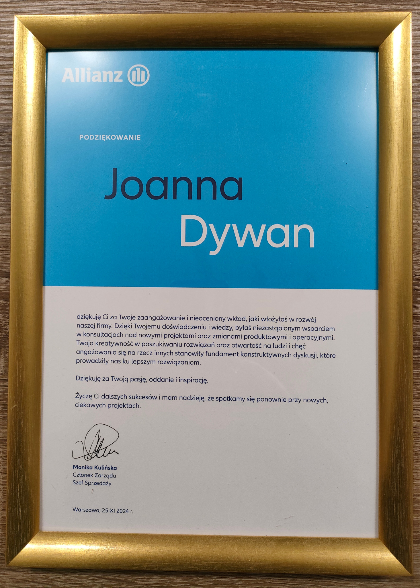 Oprawione w złotą ramę podziękowanie od Allianz dla Joanny Dywan za wkład w rozwój firmy, z podpisem Moniki Kulińskiej, datowane na 25.11.2024.