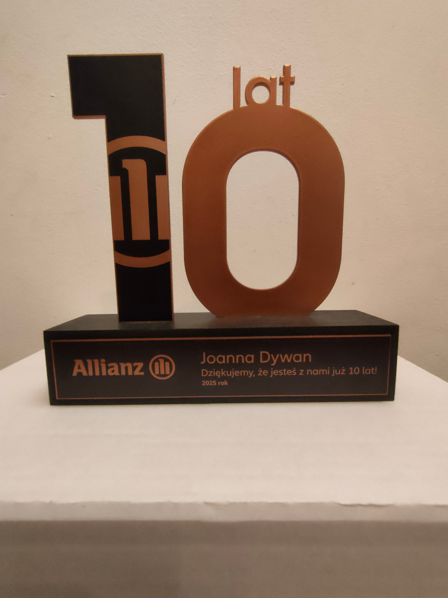 Statuetka z logo Allianz, upamiętniająca 10-lecie pracy Joanny Dywan, z napisem 'Dziękujemy, że jesteś z nami już 10 lat! 2025 rok'.