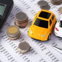 Kalkulator, monety, zabawkowy ż&oacute;łty i biały samoch&oacute;d oraz srebrny długopis na arkuszu kalkulacyjnym z danymi finansowymi.