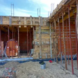 Budowa ściany z czerwonej cegły z widocznym szalunkiem drewnianym i podporami pionowymi, na pierwszym planie piasek i wiadro