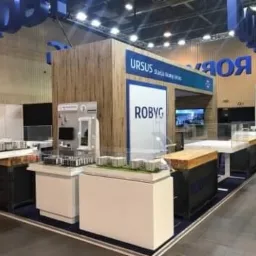 Stoisko targowe firmy Ursus Robyg z modelami osiedli mieszkaniowych i panelem prezentującym wyposażenie łazienki na tle drewnianej ściany z logo.