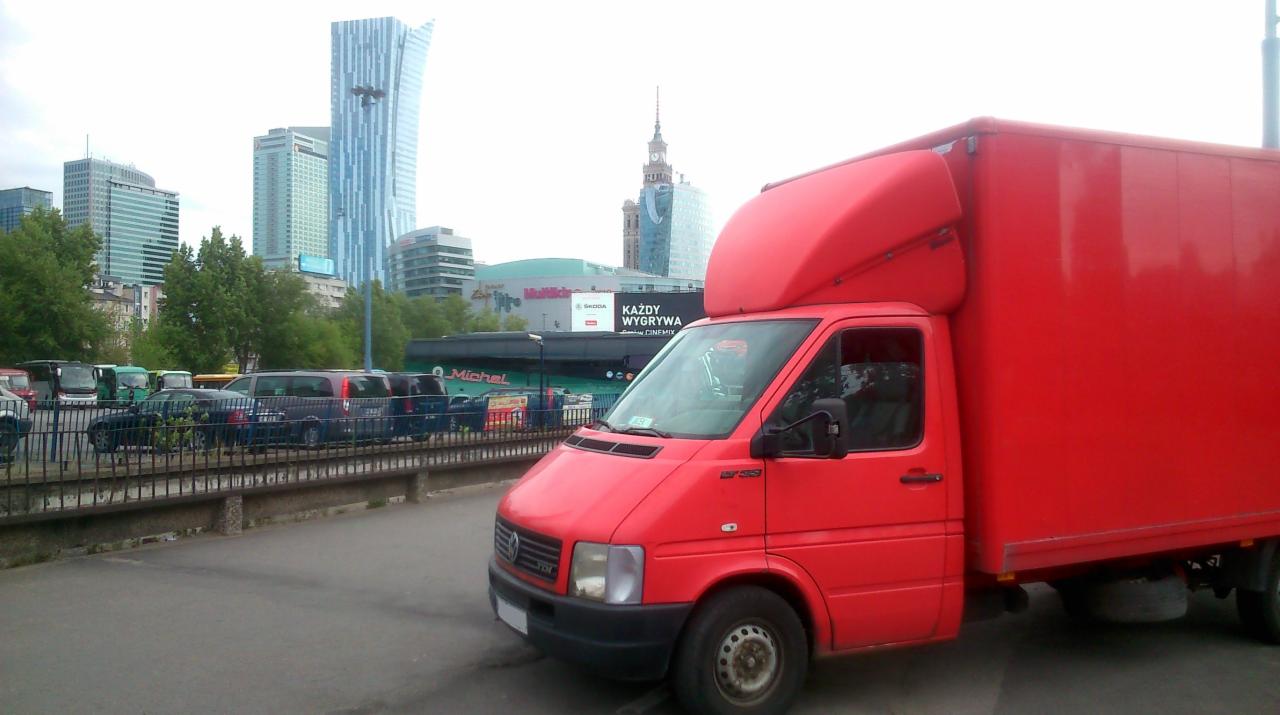 Czerwony bus dostawczy na parkingu miejskim z widokiem na Pałac Kultury i Nauki w Warszawie. Pojazd zaparkowany obok innych busów i samochodów osobowych.