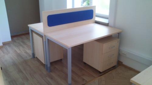 Dwa stanowiska pracy biurowej z jasnymi blatami i szarymi nogami, oddzielone pionową przegrodą z niebieskim panelem akustycznym, z szafkami kontenerowymi pod blatami, na podłodze panele imitujące...