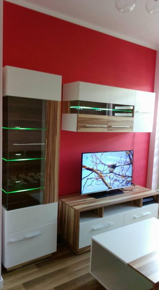 Nowoczesny zestaw mebli do salonu z białymi frontami i drewnianymi akcentami, podświetlane półki w witrynie, czerwona ściana w tle, telewizor z widokiem zimowego krajobrazu.