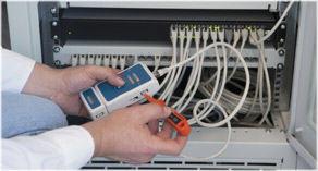 Technik testuje kable sieciowe w szafie rack za pomocą testera okablowania. Widoczne porty patch panelu z podłączonymi kablami oraz narzędzie do testowania połączeń.