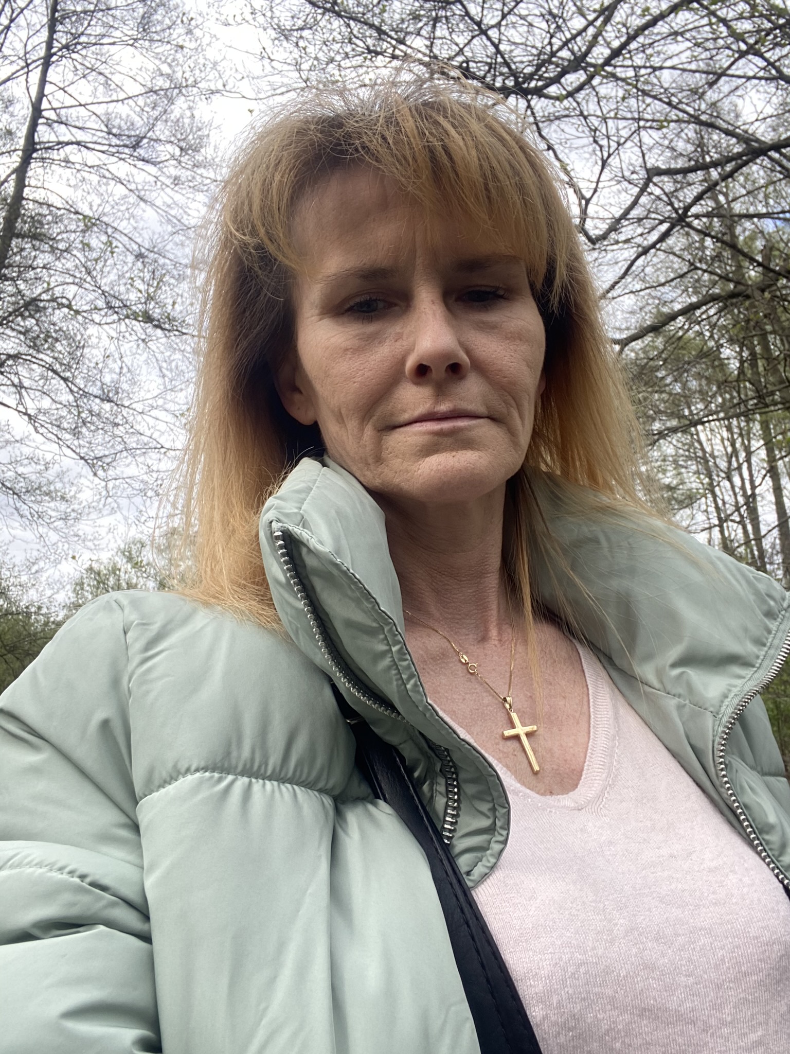 Portret kobiety w miętowej kurtce puchowej i złotym naszyjniku z krzyżykiem na tle drzew bez liści.