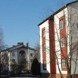 Dwa budynki mieszkalne, jeden z czerwoną elewacją i białymi pasami, drugi szary z elementami dekoracyjnymi. Na pierwszym planie drzewa bez liści i zaparkowane samochody.