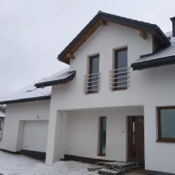 Nowoczesny, biały dom z dwuspadowym dachem pokrytym czarną dachówką, z balkonami zabezpieczonymi metalowymi barierkami, widziany w zimowej scenerii podczas opadów śniegu.