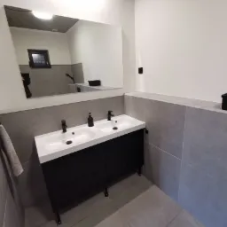 Nowoczesna łazienka z podwójną białą umywalką na czarnej szafce, szare płytki ścienne i podłogowe, duże lustro z odbiciem okna i czarnego pojemnika.