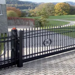 Otwarta, przesuwna brama wjazdowa z czarnego metalu z dekoracyjnym elementem w kształcie okręgu z ornamentem, na podjeździe z kostki brukowej, w tle dom z kamienia i zielony krajobraz.