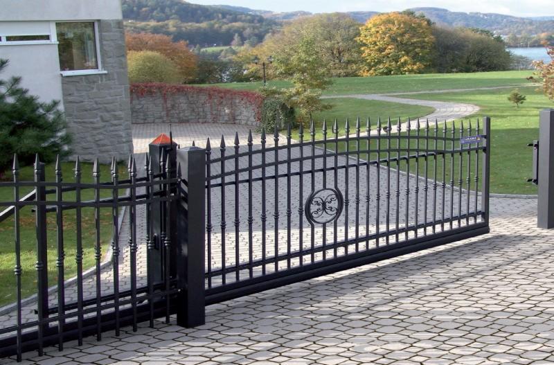 Otwarta, przesuwna brama wjazdowa z czarnego metalu z dekoracyjnym elementem w kształcie okręgu z ornamentem, na podjeździe z kostki brukowej, w tle dom z kamienia i zielony krajobraz.