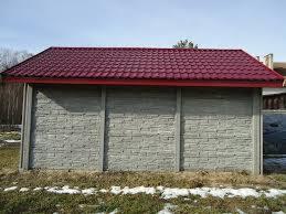 Betonowa wiata z czerwonym dachem z blachodachówki, widoczna na tle drzew i resztek śniegu na trawie.