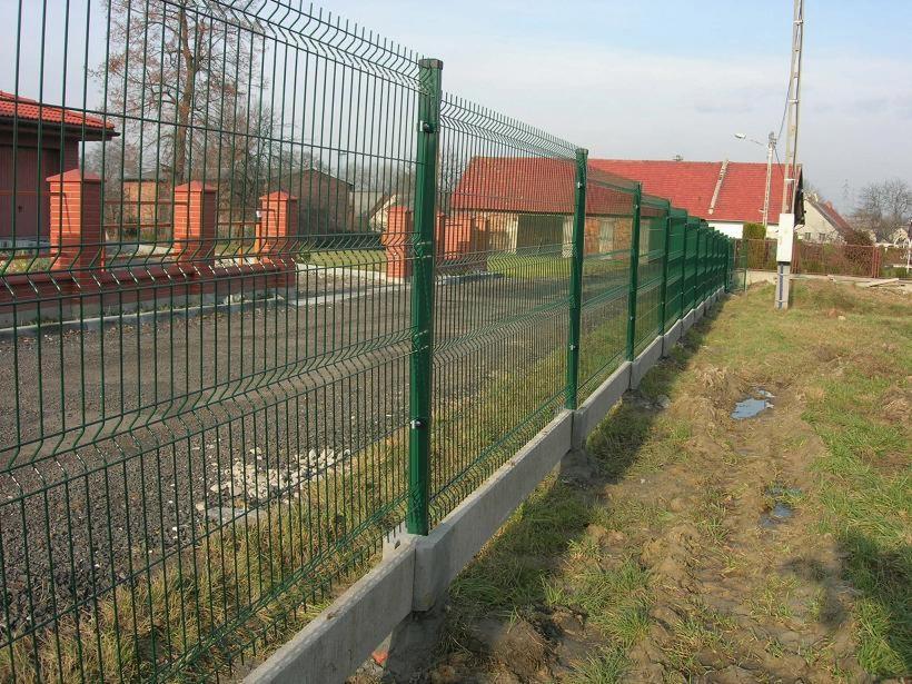 Długi, zielony panelowy płot z betonową podmurówką, widziany z boku na tle domów z czerwonymi dachami, w słoneczny dzień. Widoczne ślady opon na trawie.