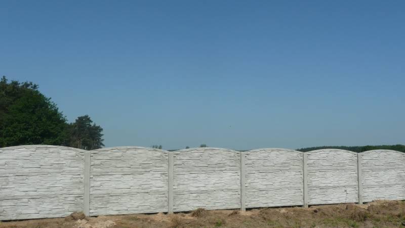 Gotowy, betonowy płot imitujący kamień łupany z zaokrąglonymi górnymi krawędziami paneli, na tle błękitnego nieba i zieleni drzew