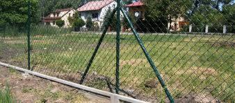 Zielona siatka ogrodzeniowa na betonowej podmurówce, widoczne podpory ukośne, w tle domy jednorodzinne i drzewa.