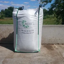 Worek nawozu naturalnego marki Bionics z zielonymi uchwytami, umieszczony na betonowej powierzchni z polami i drzewami w tle.