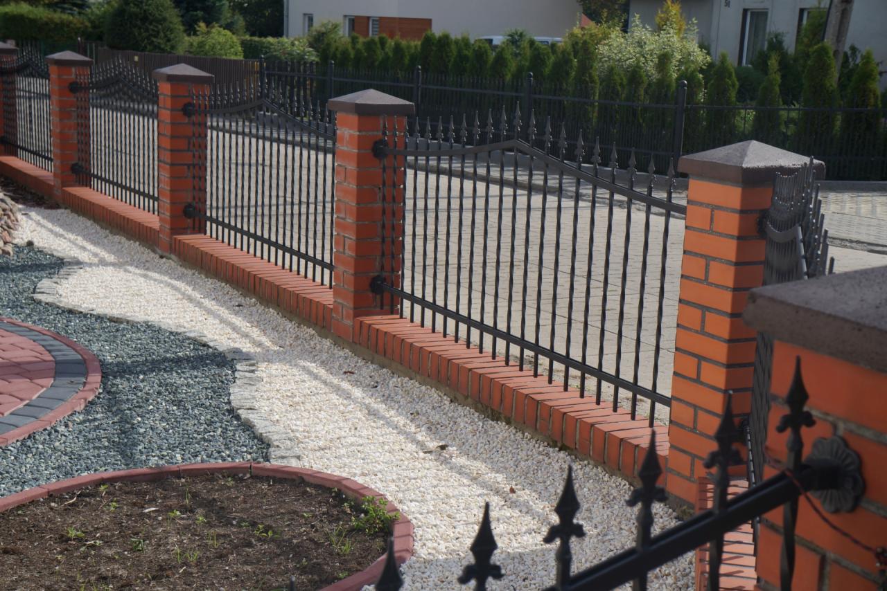 Kute, czarne ogrodzenie z ceglanymi słupkami i podmurówką, otoczone ozdobnym, białym i szarym żwirem oraz fragmentem trawnika z obrzeżem z kostki.