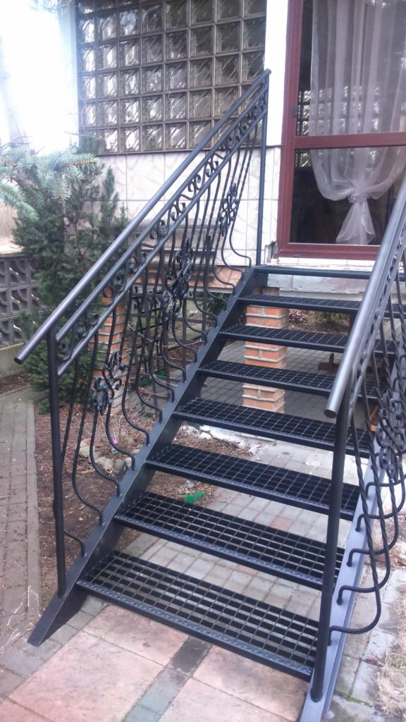 Zewnętrzne, metalowe schody kratowe z ozdobnymi poręczami prowadzące do drzwi wejściowych budynku.