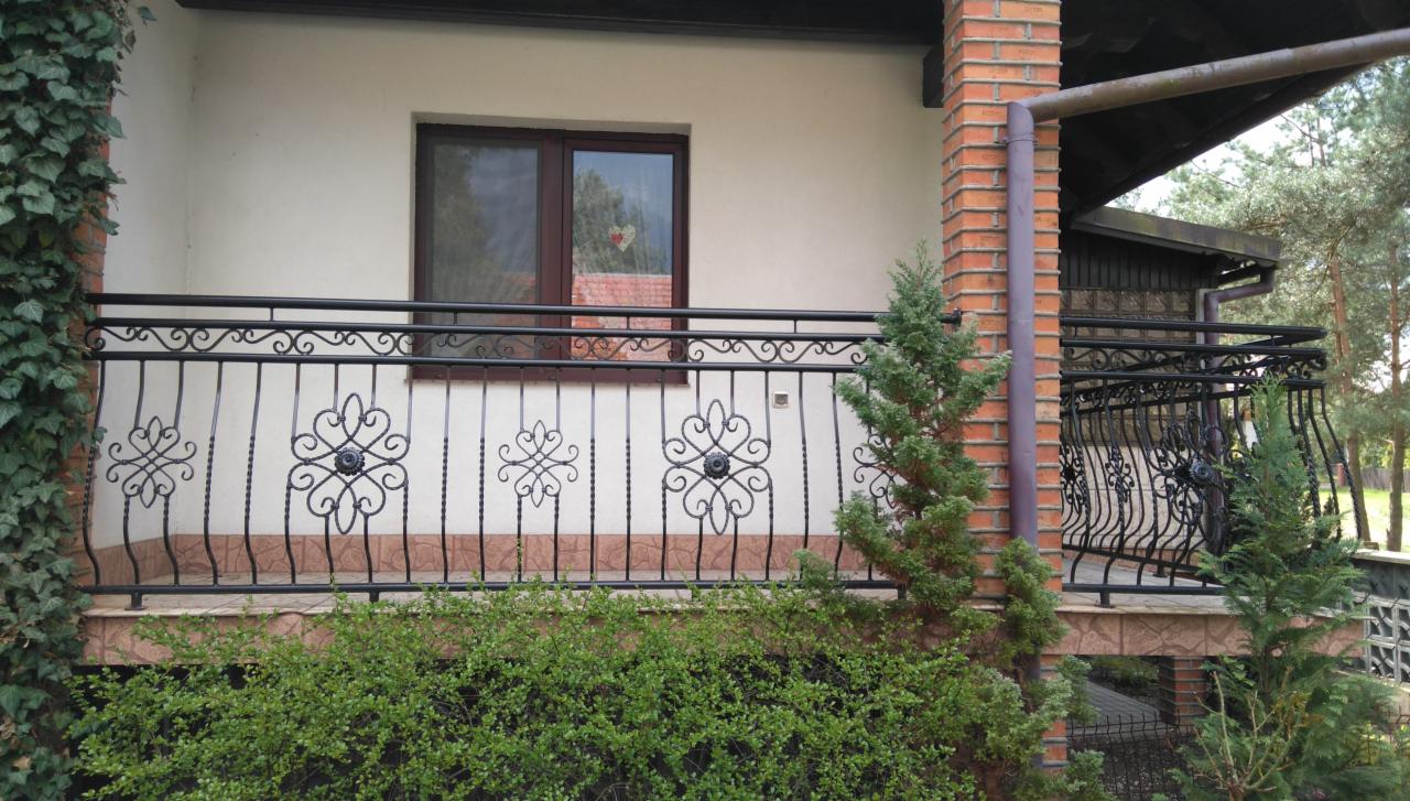 Kuty balkon z ozdobnymi elementami, widok z zewnątrz, z roślinnością w tle i na pierwszym planie.