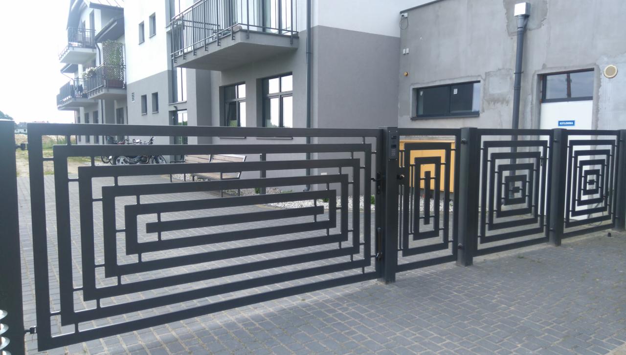 Nowoczesna, metalowa brama posesyjna z geometrycznym wzorem labiryntu, widok z zewnątrz, na tle bloku mieszkalnego z balkonami.