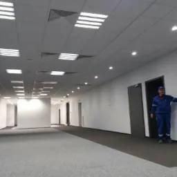 Długi korytarz biurowy z równomiernie rozmieszczonymi panelami oświetleniowymi na suficie podwieszanym, pracownik w niebieskim kombinezonie wskazuje na ścianę.