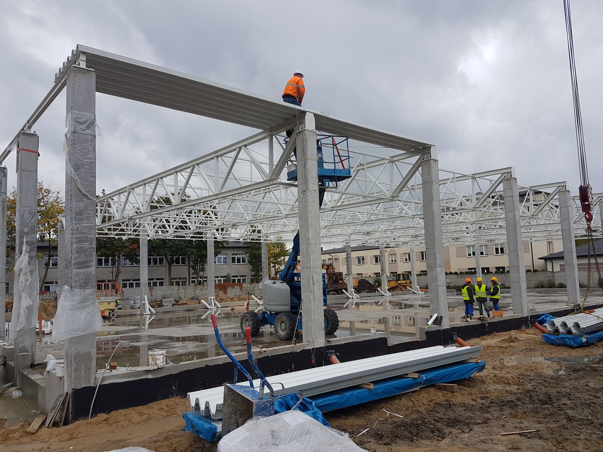 Konstrukcja stalowa hali przemysłowej w trakcie montażu, pracownik w pomarańczowej kurtce na podnośniku, betonowe słupy zabezpieczone folią, w tle budynki mieszkalne.