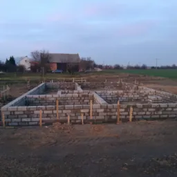 Wznoszone fundamenty domu z bloczków betonowych, widoczne drewniane podpory stabilizujące, w tle budynki gospodarcze i pole, pochmurne niebo.