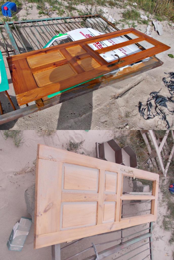 Dwie drewniane drzwi z szybami, górne po renowacji z brązową bejcą, dolne surowe, ułożone na metalowych kratach na piaszczystym podłożu. Widok z góry.