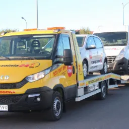 Ż&oacute;łta laweta Iveco z napisem 'Pomoc Drogowa' i numerem telefonu przewozi dwa samochody osobowe na przyczepie. Widok z przodu, oświetlenie dzienne.