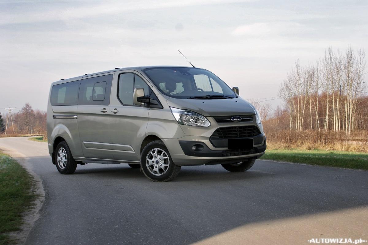 Szaro-beżowy Ford Transit Custom na asfalcie, z anteną na dachu, na tle drzew i nieba. Widok z przodu, lekko z boku, podkreślający przestronność wnętrza.