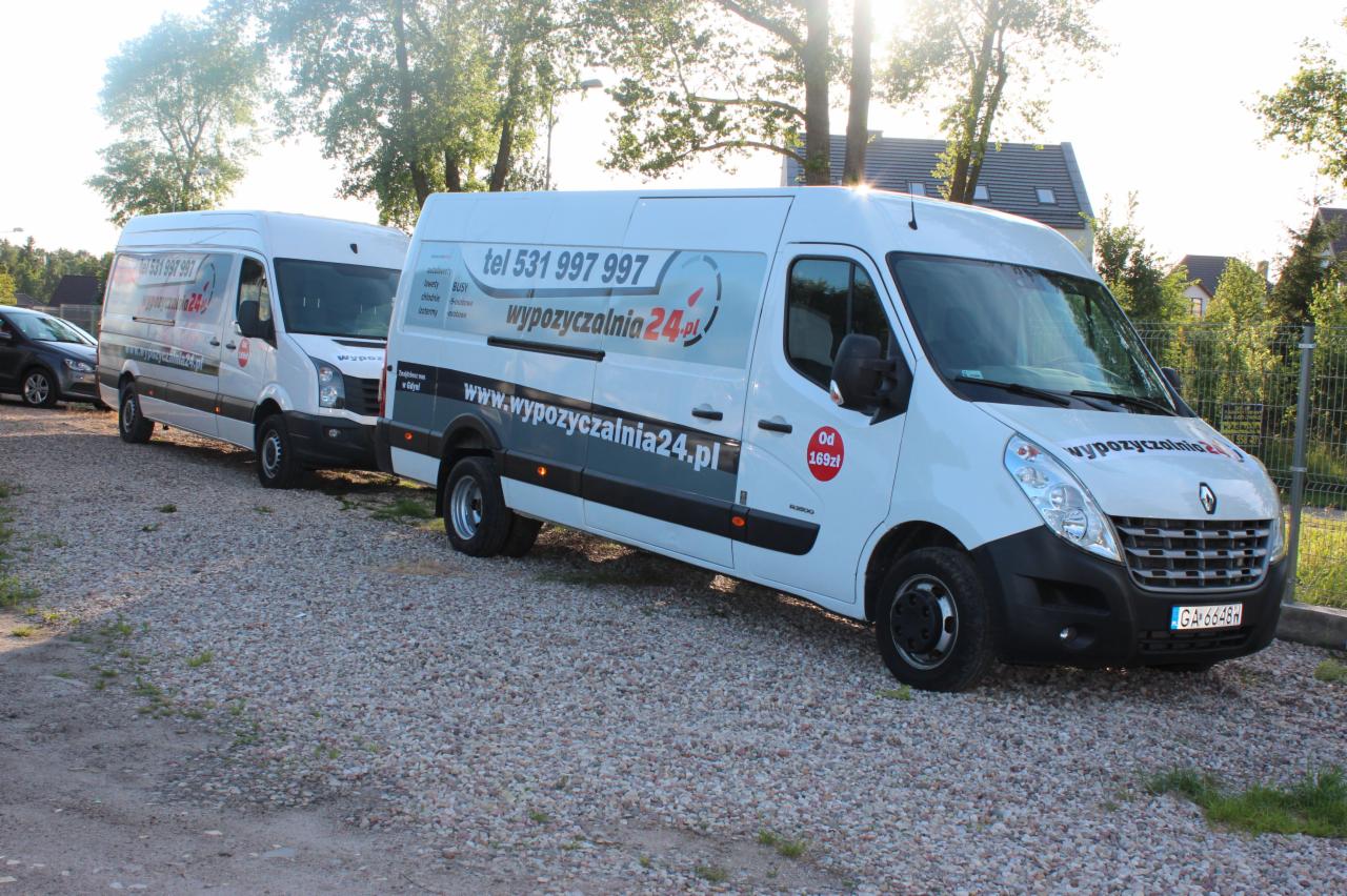 Dwa białe busy z logo wypożyczalni na żwirowym parkingu w słoneczny dzień. Na pierwszym planie widoczny przód busa z numerem rejestracyjnym.