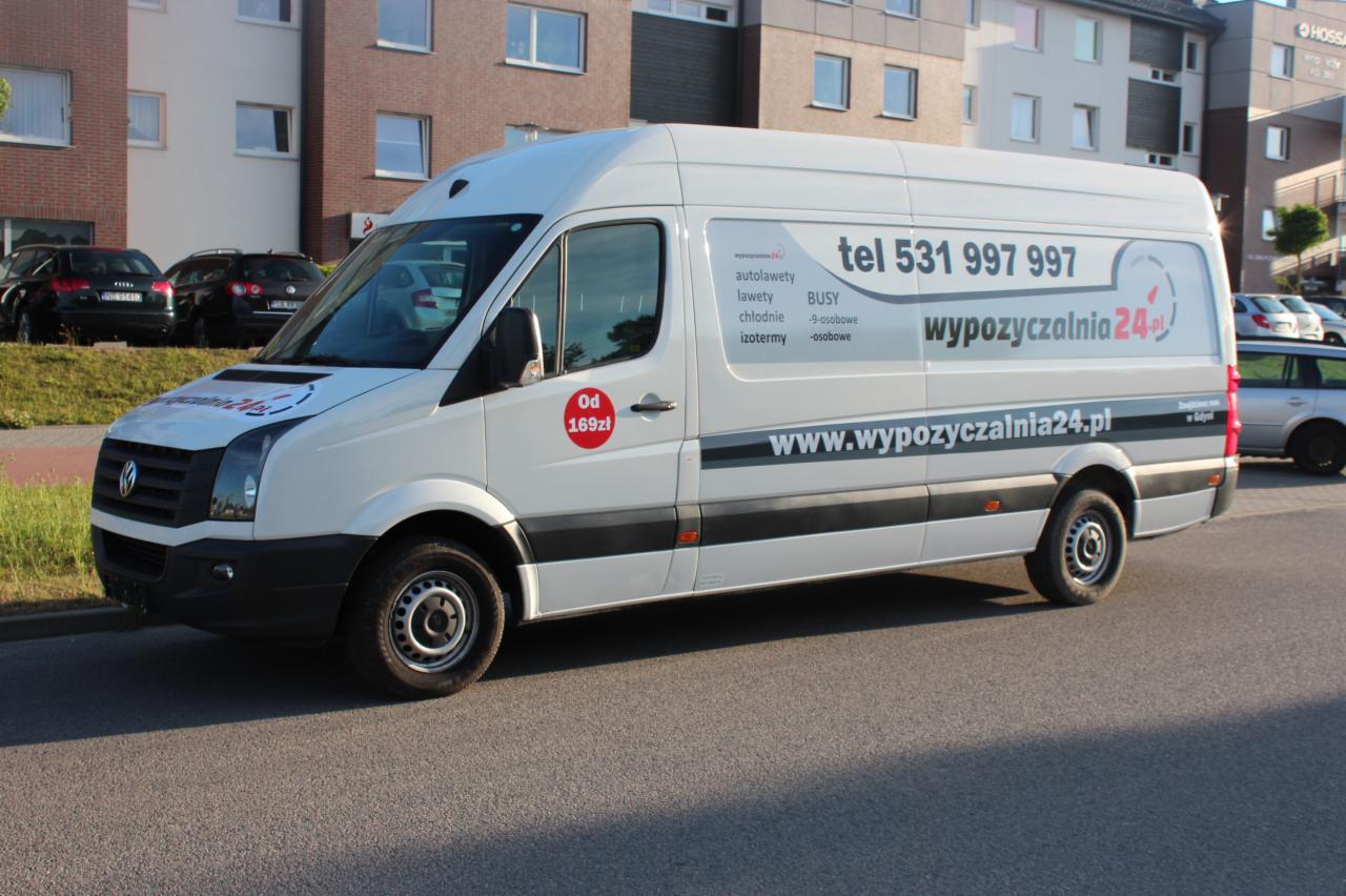 Biały bus z reklamą wypożyczalni autolawet, lawet, busów chłodni i izotermicznych, zaparkowany na ulicy przed budynkiem. Widoczny numer telefonu i adres strony internetowej.