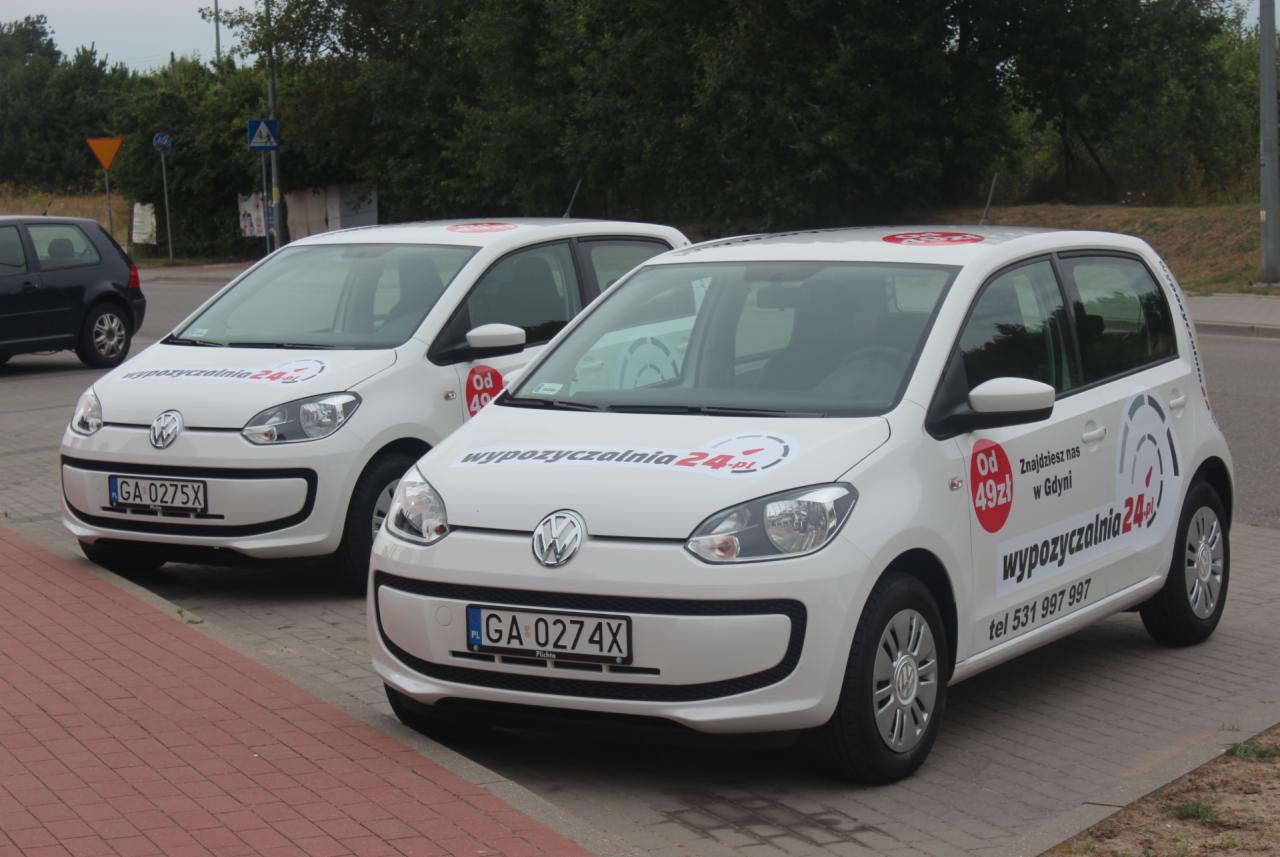 Dwa białe Volkswageny Up z logo wypożyczalni samochodów na parkingu. Auta z tablicami GA 0275X i GA 0274X, reklama 'Od 49 zł' i numer telefonu.