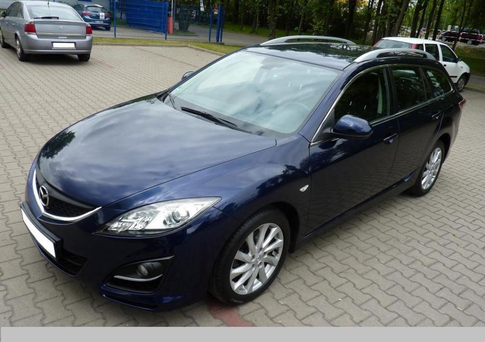 Granatowa Mazda 6 kombi, widok z przodu i boku, zaparkowana na kostce brukowej. Widoczne srebrne relingi dachowe, alufelgi i inne pojazdy w tle.