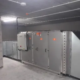 Szara centrala wentylacyjna z panelem sterowania, widoczna w surowym pomieszczeniu z betonowym sufitem i ścianami z bloków, oświetlona lampą sufitową.