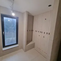 Ściana łazienki w trakcie układania płytek z użyciem klipsów poziomujących, widoczne pionowe wzory na płytkach i okno w czarnej ramie.