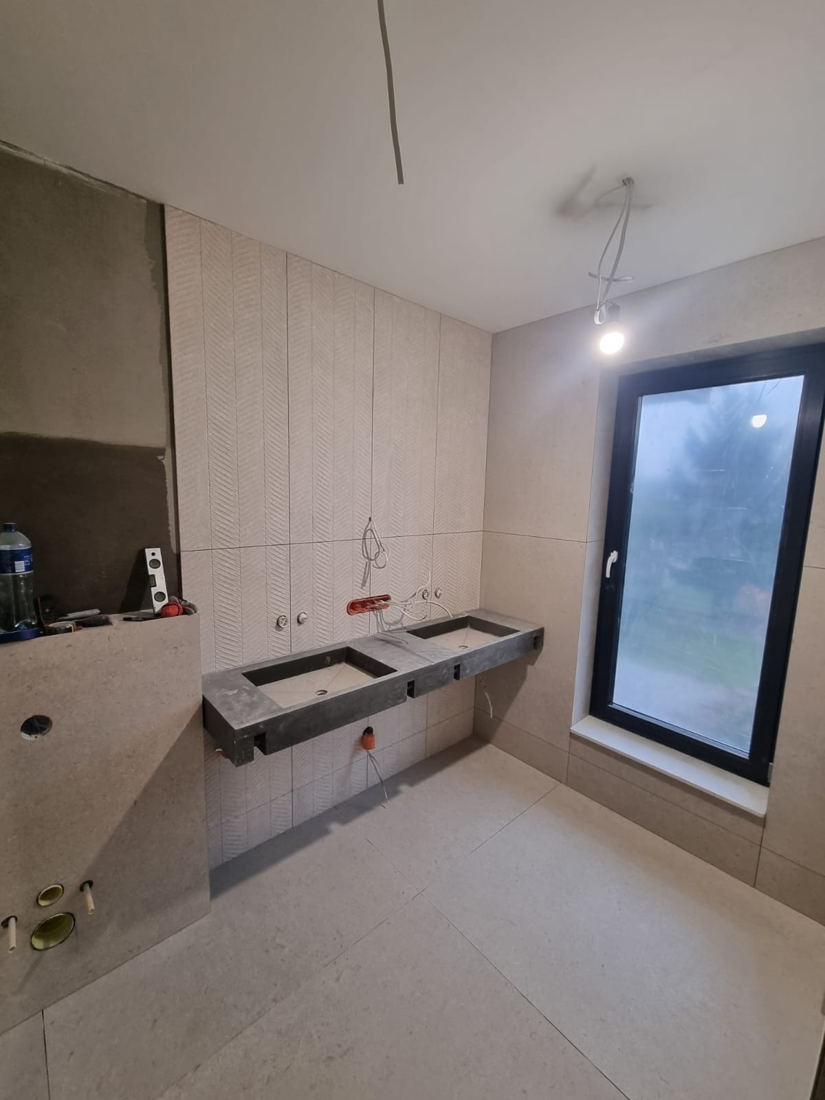 Łazienka w trakcie prac wykończeniowych; widoczne dwie szare umywalki podwieszane, płytki ścienne ułożone we wzór chevron, okno z czarną ramą i niedokończone instalacje elektryczne.