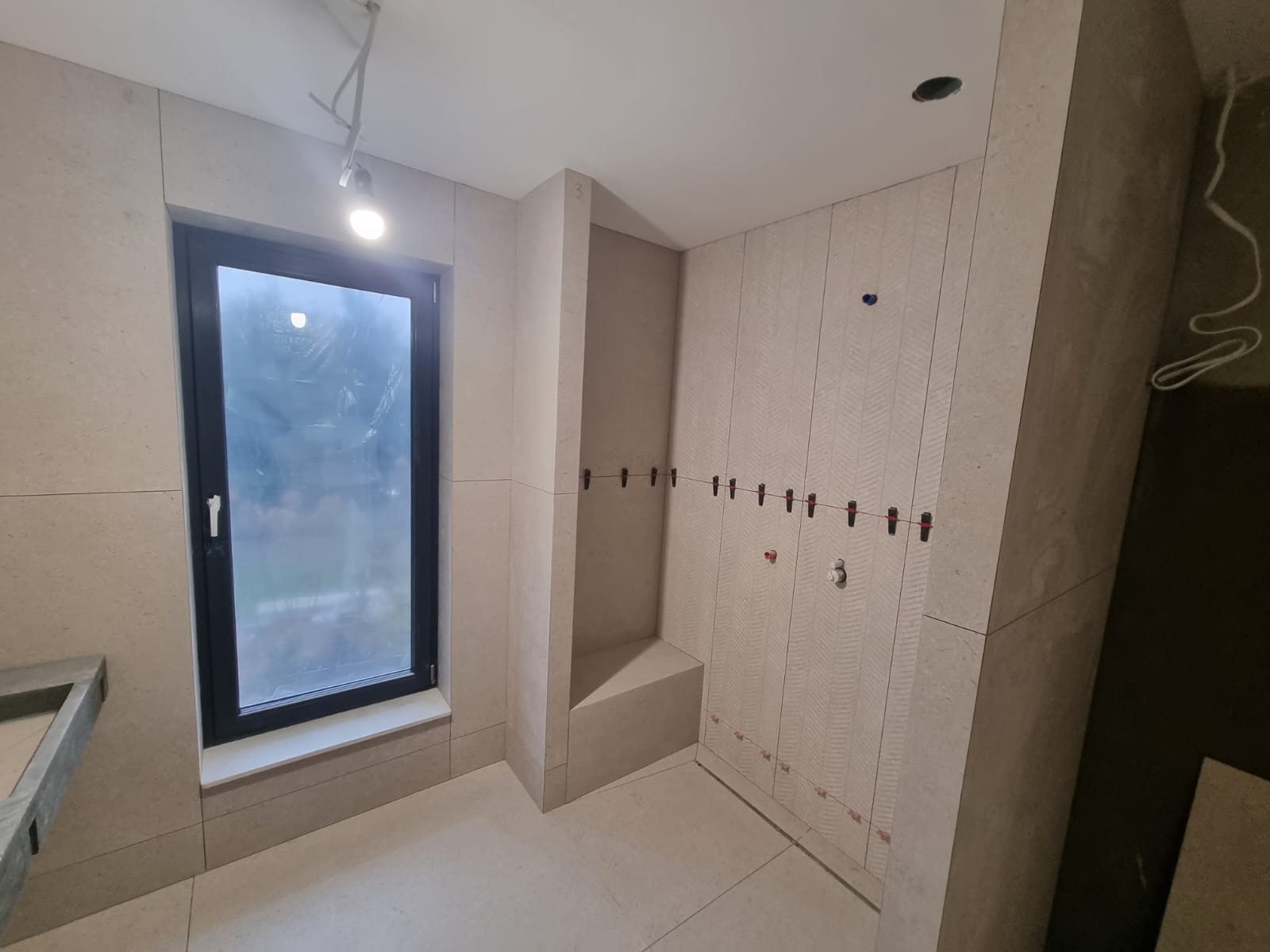 Ściana łazienki w trakcie układania płytek z użyciem klipsów poziomujących, widoczne pionowe wzory na płytkach i okno w czarnej ramie.