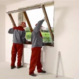 Dwóch monterów w czerwono-szarych uniformach instaluje okno PCV w surowym stanie, widoczny poziomica przy ścianie i grzejnik pod oknem.