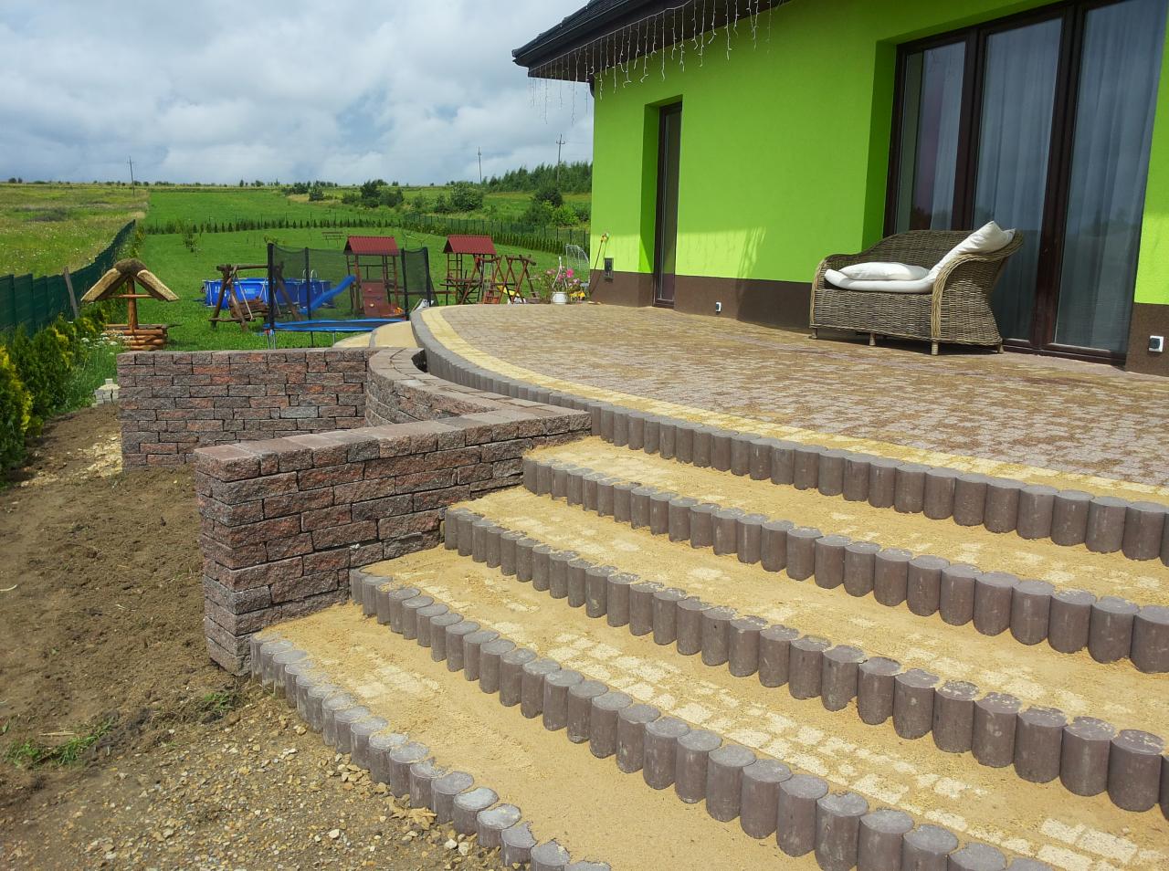 Schody z obrzeżami i piaskiem prowadzą na taras z kostki brukowej. W tle zielony budynek i krajobraz z placem zabaw.